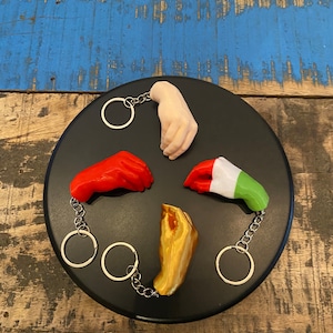 Peut inclure: Cinq porte-cl&eacute;s en forme de main sur un fond noir. Les porte-cl&eacute;s sont faits de diff&eacute;rents mat&eacute;riaux, dont le plastique, le m&eacute;tal et le bois. Les porte-cl&eacute;s sont de diff&eacute;rentes couleurs, notamment rouge, blanc, vert, or et couleur chair.