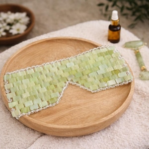 May include: A light green jade eye mask with a white trim, resting on a wooden plate. The mask is made of small, rectangular jade stones. A small bottle and a jade roller are also visible.