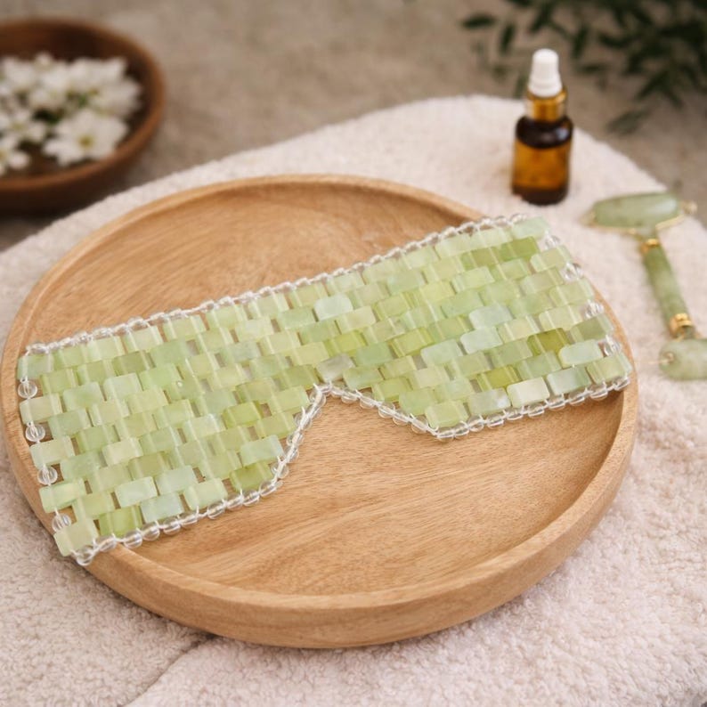 May include: A light green jade eye mask with a white trim, resting on a wooden plate. The mask is made of small, rectangular jade stones. A small bottle and a jade roller are also visible.