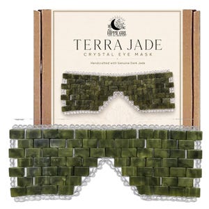 Op de afbeelding: Een donkergroen jade kristallen oogmasker bestaande uit rechthoekige stenen tegels verbonden door doorzichtige kralen. Het masker wordt getoond voor een kartonnen doos met het label "Terra Jade Crystal Eye Mask" en "Handcrafted with Genuine Dark Jade."