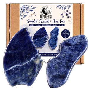 May include: Two blue sodalite stone facial tools in front of a cardboard box. The box features the text "Sodalite Sculpt + Flow Duo", "MADE FROM A+ GRADE BLUE SODALITE STONE", and "FACIAL SCULPTING & SELF-CARE" with a logo showing a moon and crystals.