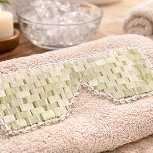 May include: A light green jade eye mask with a woven design, resting on a beige towel. The mask is surrounded by a clear beaded trim. A lit candle, a bowl of ice, and a rolled towel are in the background.