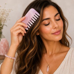 May include: A light pink rose quartz comb is used on long, wavy brown hair. A rose quartz bracelet with a gold charm and a gold necklace with a rose quartz pendant are also visible. The comb has a rectangular shape with evenly spaced teeth.