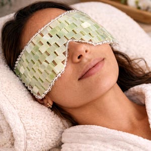 May include: A woman wearing a light green jade eye mask with a woven design, resting on a white towel. The mask is trimmed with a white beaded border. The woman is wearing a white robe.