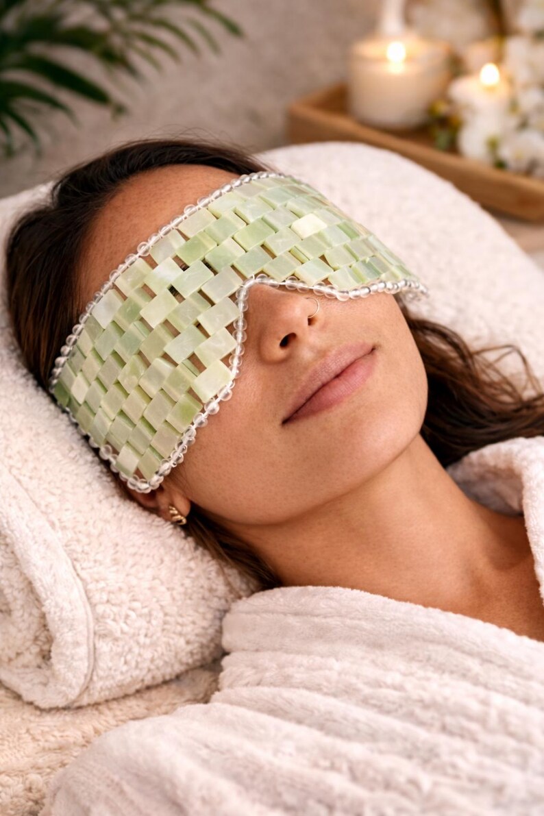 May include: A woman wearing a light green jade eye mask with a woven design, resting on a white towel. The mask is trimmed with a white beaded border. The woman is wearing a white robe.