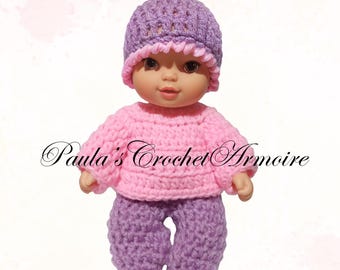 May include: A doll wearing a handmade crochet outfit. The outfit includes a pink sweater, purple pants, and a purple hat with a pink brim and pom-pom. The text "Paula's Crochet Armoire" is visible.