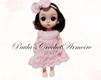 May include: A doll wearing a light pink crocheted dress and matching bow headband. The doll has dark hair and large eyes. The text "Paula's Crochet Armoire" is written in cursive below the doll.