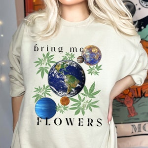 May include: Cream-colored sweatshirt with a graphic design featuring the Earth, planets, and cannabis leaves. The text "bring me FLOWERS" is printed in black. The design has a celestial and botanical theme.