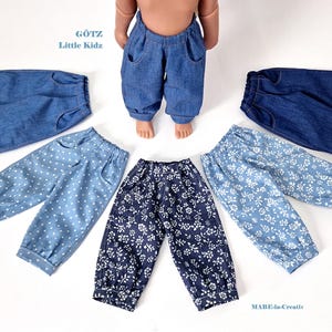 May include: Doll trousers in a variety of denim and patterned fabrics. The collection includes solid blue denim, polka dot, and floral designs. The image includes the text "Götz Little Kidz" and "MABE-in-Creativ".