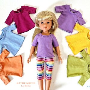 Könnte beinhalten: Ein Set mit 7 bunten T-Shirts in verschiedenen Farben wie Lila, Gelb, Grün, Orange, Blau und Pink für Puppen. Die Shirts liegen um eine Puppe herum, die ein lila T-Shirt und regenbogenfarbene Leggings trägt. Die Puppe ist ein blondes Mädchen mit blauen Augen. Der Text "Käthe Kruse La Bella" ist am unteren Rand des Bildes zu sehen.