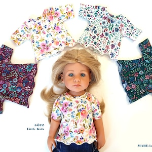 Op de afbeelding: Vijf poppenblouses met bloemenprint in verschillende kleuren, waaronder wit, groen, bordeauxrood en roze. De blouses zijn gerangschikt rond een pop die een vergelijkbare blouse met bloemenprint draagt. De tekst "GÖTZ Little Kidz" is zichtbaar op de afbeelding.