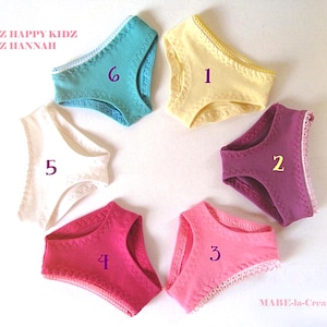 Könnte beinhalten: Sechs Paar Puppenunterhosen, die in einem Kreis angeordnet und mit den Zahlen eins bis sechs beschriftet sind. Die Farben umfassen Gelb, Lila, Pink, Dunkelpink, Weiß und Türkis. Jedes Paar hat eine dekorative Zickzacknaht an den Rändern. Der Text lautet GÖTZ HAPPY KIDZ GÖTZ HANNAH und MABE-la-Creativ.