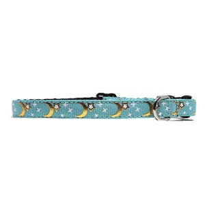 May include: A light blue cat collar with a repeating pattern of gold crescent moons and white stars. The collar has a black plastic buckle and a silver metal D-ring for attaching a leash.