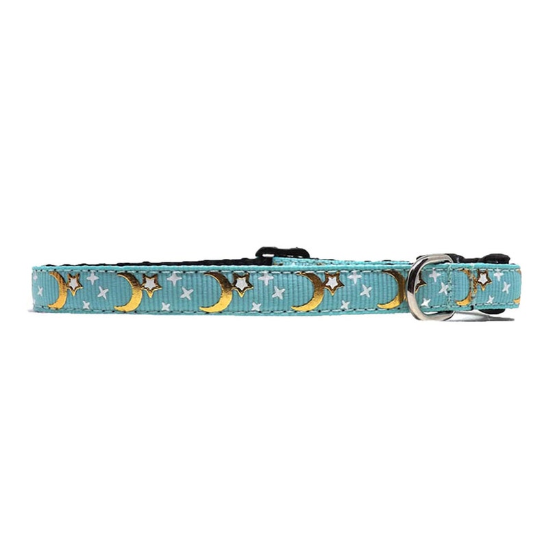 May include: A light blue cat collar with a repeating pattern of gold crescent moons and white stars. The collar has a black plastic buckle and a silver metal D-ring for attaching a leash.