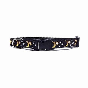 May include: A black pet collar featuring a repeating pattern of metallic gold crescent moons and white stars. The collar includes a black plastic side-release buckle, a silver-toned metal D-ring for lead attachment, and a black plastic sliding adjuster.