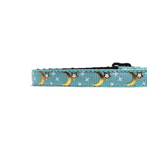 May include: A light blue pet collar with a repeating pattern of gold crescent moons and white stars. The collar has a black buckle and is designed for a small pet. The celestial design adds a touch of whimsy.