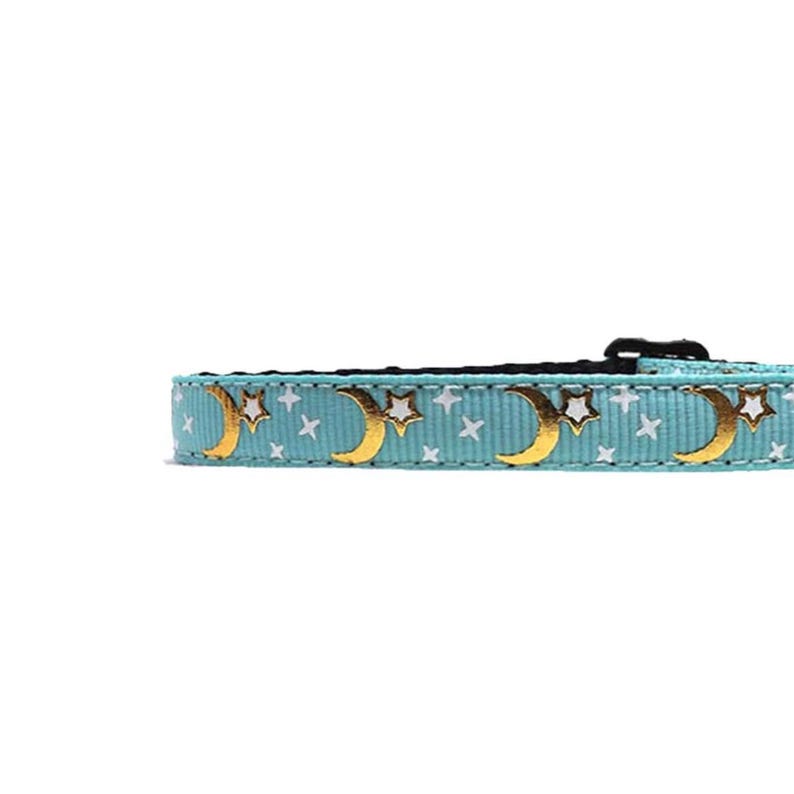 May include: A light blue pet collar with a repeating pattern of gold crescent moons and white stars. The collar has a black buckle and is designed for a small pet. The celestial design adds a touch of whimsy.