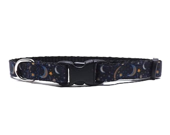 May include: A black pet collar featuring a celestial pattern of yellow stars, crescent moons, and small planets. The collar includes a black plastic side-release buckle, a silver-toned metal D-ring for leash attachment, and a black plastic sliding adjuster.