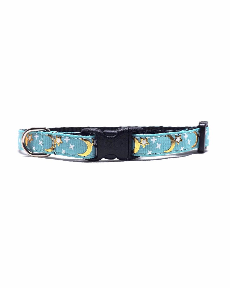 May include: A light blue pet collar with a repeating pattern of gold moons and white stars. The collar has a black plastic buckle and a silver metal D-ring for leash attachment.