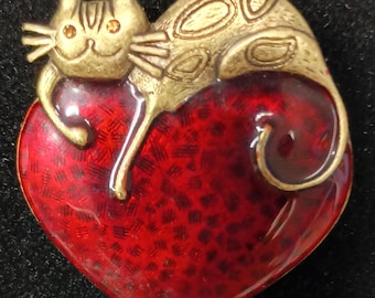 May include: A gold-toned metal brooch shaped like a cat resting on a large heart. The cat has small orange gemstone eyes and textured markings on its body. The heart is finished with a deep red enamel featuring a dark, mottled, and textured pattern.