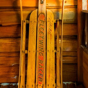 May include: A vintage wooden sled with a black metal frame and the words "Fleetwing  Sled" painted on the side. The sled is leaning against a wooden wall.