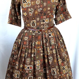 May include: A brown collared dress featuring a repeating grid pattern of various fruits, flowers, and leaves in shades of orange, yellow, green, and black. The dress has short sleeves, a button-down front, and a thin brown belt at the waist.