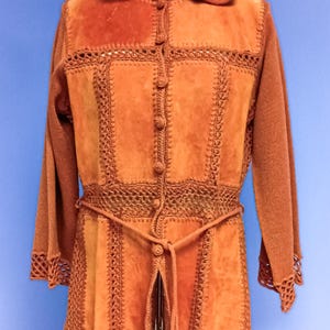 Può includere: Giacca in camoscio color ruggine con cappuccio, maniche lunghe e cintura all'uncinetto. La giacca presenta una chiusura con bottoni sul davanti e dettagli all'uncinetto lungo le cuciture e i polsini. La giacca è esposta su un manichino.