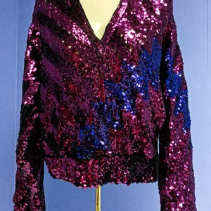 May include: A sequinned V-neck sweater in shades of purple, black, and blue. The long-sleeved top features a ribbed hem and cuffs. The sequins create a shimmering effect, with a pattern of stripes and a blue design on the front.