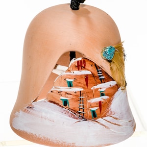 May include: A terracotta bell with a painted scene of a pueblo-style building in a snowy landscape. The bell has a black rope handle and a turquoise stone accent.