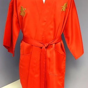 May include: Bright orange satin robe with a matching belt, displayed on a mannequin. The robe features two pockets and gold embroidered symbols near the shoulders. The robe is a V-neck style.
