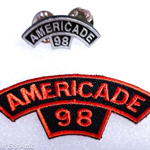 May include: Two items: a pin and a patch. The pin is silver with the word "AMERICADE" arched across the top and the number "98" below. The patch is black with red stitching and the word "AMERICADE" arched across the top and the number "98" below.