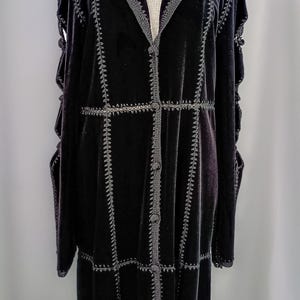 May include: A long, black velvet coat with intricate gray stitching details. The coat features a notched collar, button closures, and decorative seams. The sleeves have a unique open design. The coat is displayed on a mannequin.