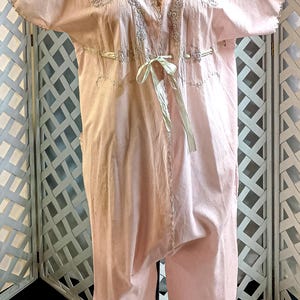 May include: A light pink vintage romper with short sleeves and a V-neckline. The romper features embroidered detailing around the neckline and sleeves, with a ribbon tie at the waist. The bottom portion is a loose fit.