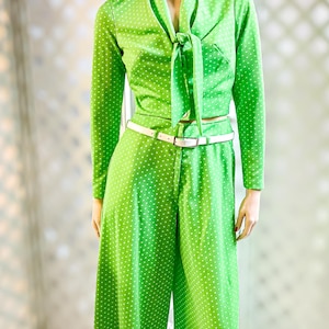 Puede incluir: Un pantalón de lunares verdes vintage en un maniquí. El conjunto incluye una blusa de manga larga con lazo en el cuello, pantalones de pernera ancha y un cinturón blanco. El maniquí tiene una peluca rizada marrón y pendientes verdes.
