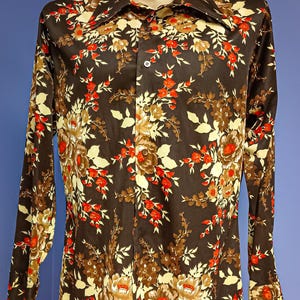 May include: A long-sleeved button-down shirt with a dark brown background and a floral pattern in red, yellow, and brown. The shirt has a wide collar and buttoned cuffs. The text "Aunt B's Attic" is visible at the bottom.