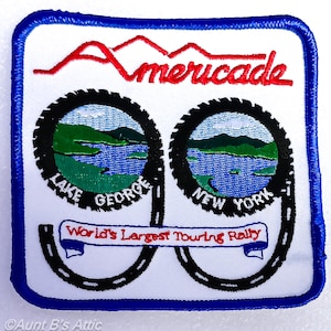 May include: A square patch with a white background and a blue border. The patch features the word "Americade" in red, above two black tire-shaped designs with lake and mountain scenes. The text "Lake George New York" is included. The words "World's Largest Touring Rally" are also present.