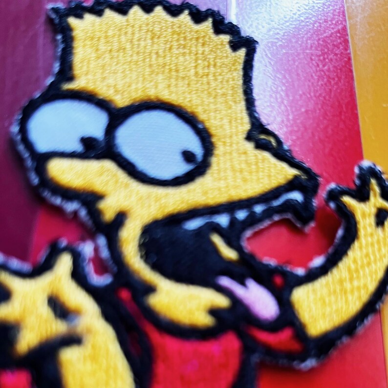 K&ouml;nnte beinhalten: Gestickter Aufn&auml;her mit Bart Simpson, gelben Haaren, blauen Augen und einem roten Hemd. Der Aufn&auml;her hat einen schwarzen Rand und zeigt Bart mit herausgestreckter Zunge. Der Hintergrund ist eine Mischung aus Rot, Kastanienbraun und Gelb.