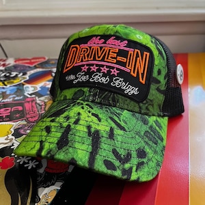 May include: A green and black trucker hat with a camouflage pattern. The front patch reads "The Last Drive-In with Joe Bob Briggs" in orange and pink lettering. The hat has a black mesh back and a white button.