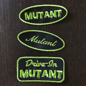 May include: Three black fabric patches with green embroidered text. The top patch reads "MUTANT" in block letters. The middle patch reads "Mutant" in cursive. The bottom patch reads "Drive-In MUTANT" in block letters.