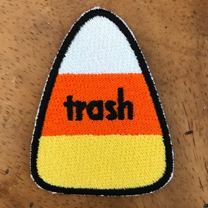 Peut inclure: Patch en forme de maïs sucré orange, jaune et blanc avec un contour noir et le mot "trash" brodé en noir.