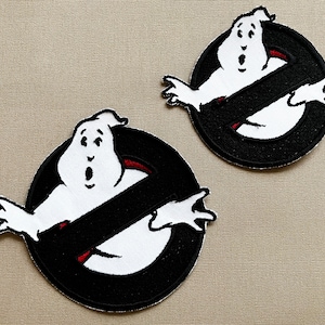 May include: Two black and white embroidered patches featuring the Ghostbusters logo. The logo is a circle with a diagonal line through it and a ghost inside. The ghost is white with a red mouth and eyes.