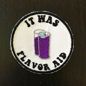 May include: A white round embroidered patch with black stitching. The patch features a purple glass of liquid with a straw and ice cubes. The text "It Has Flavor Aid" is written in a black cursive font around the glass.