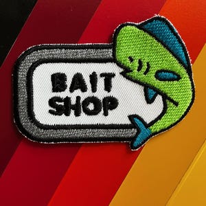 Op de afbeelding: Geborduurde patch met de tekst "BAIT SHOP" in zwarte letters in een witte rechthoek met een grijze rand. Een groene en blauwe cartoonvis bevindt zich aan de rechterkant. De achtergrond is een verloop van rood, oranje en geel.