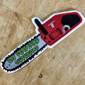 Könnte beinhalten: Roter und weißer gestickter Aufnäher einer Kettensäge mit einer grün-weißen Kette, auf der "Say Something Disturbing" steht.