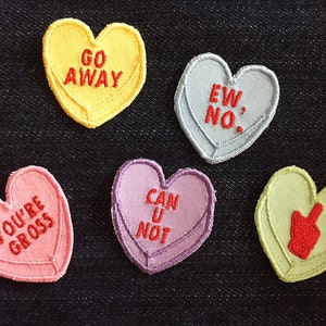 May include: Five heart-shaped patches with different messages and designs. The patches are yellow, light blue, pink, purple, and light green. The messages on the patches are "Go Away", "Ew No", "You're Gross", "Can U Not", and a middle finger.