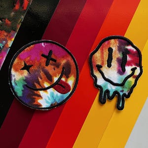 Puede incluir: Dos parches de smiley con estampado tie-dye. Uno es un smiley amarillo tradicional con bordes que gotean. El otro es un smiley tie-dye morado, naranja y azul con la lengua fuera y X para los ojos.