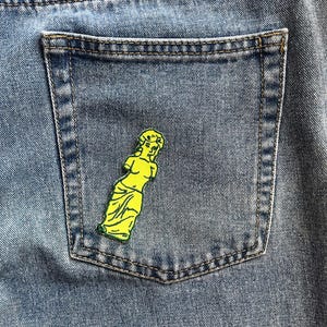 Op de afbeelding: Een gele geborduurde patch van een vrouwenfiguur met een kroon op haar hoofd, genaaid op een blauwe denim zak.