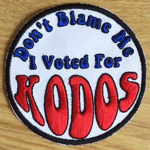 Może przedstawiać: Okrągła, biała naszywka z czarnym obramowaniem. Tekst "Don't Blame Me I Voted For KODOS" jest wyszyty na niebiesko i czerwono.