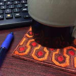 May include: A brown and orange patterned coaster with red circles, resembling the carpet from the movie "The Shining". A white mug with a black base sits on top of the coaster.