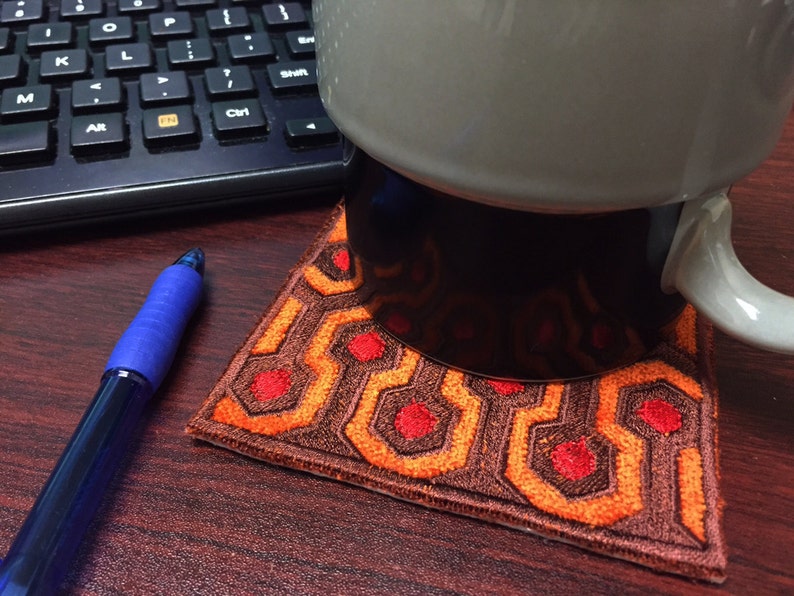 May include: A brown and orange patterned coaster with red circles, resembling the carpet from the movie "The Shining". A white mug with a black base sits on top of the coaster.